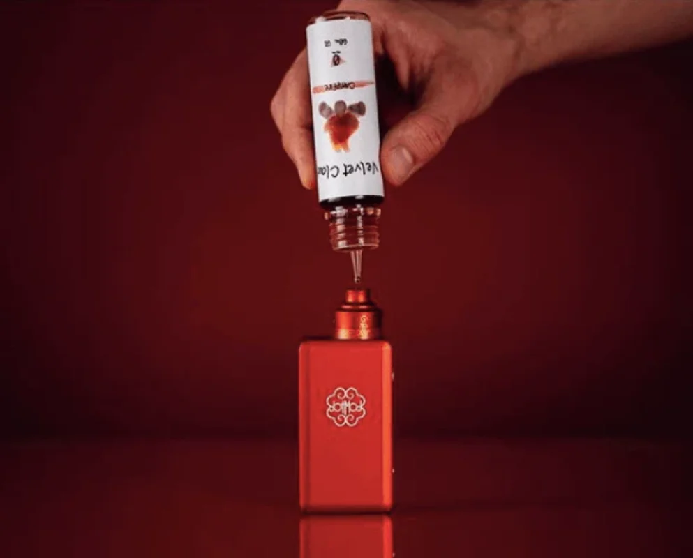 What Is a Refillable E-Cigarette and How Does It Work? 4 A hand is seen filling a red vape pod with Velvet Cola flavored e-liquid, showcasing the refill process on a red background.