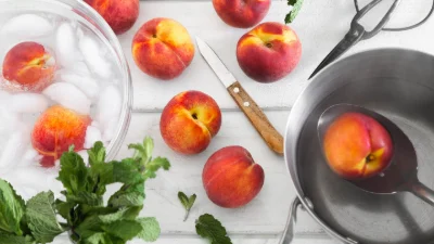 What Are the Most Popular Disposable Vape Flavors in 2026? 8 Fresh peaches on a white surface, some in an ice water bath and one being scooped from a pot with a slotted spoon. A knife and mint leaves are also visible.